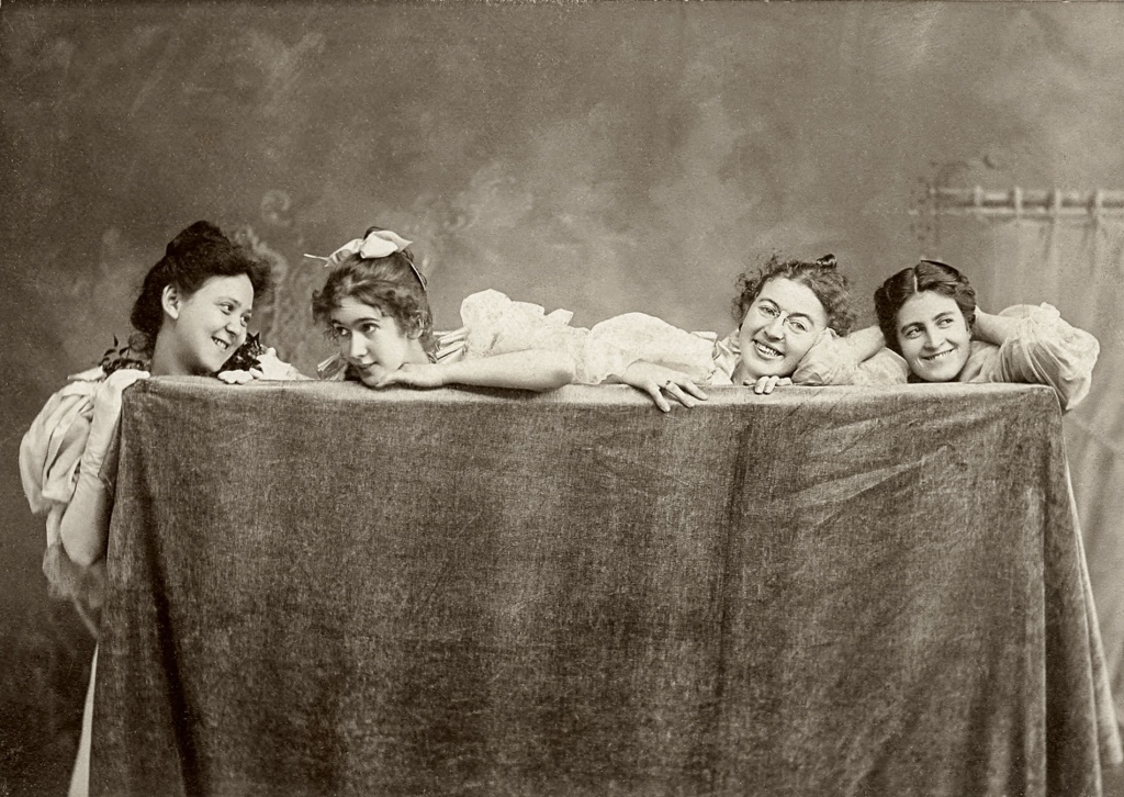 Archival image of four girls in the late 1880's leaning on a table covered with a table cloth.